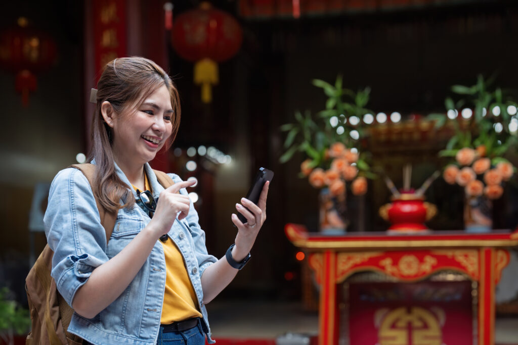 happy young asian tourist woman using smartphone on street with market background, female traveller enjoy shopping market during holidays