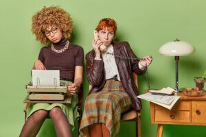 80th concept. indoor photo of two young tired african american and european women sitting in old room on green background typewriting speaking on phone at end of hard long working day in retro clothes
