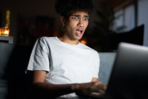 amazement. night portrait of emotional young guy looking surprised, surfing the internet, using laptop in dark room, sitting on a couch at home