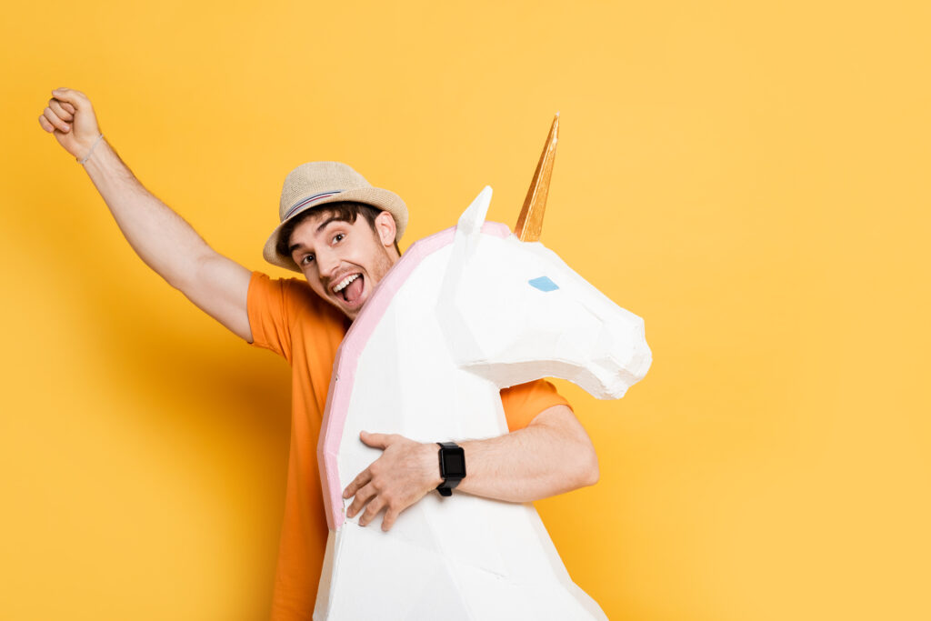 excited man in summer hat holding fist up and hugging decorative unicorn on yellow