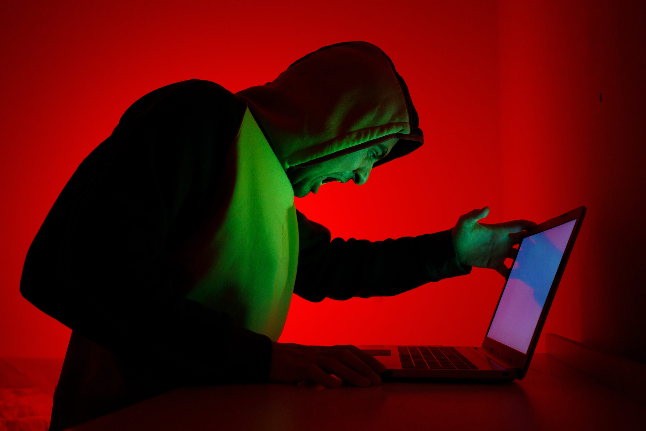 individual in hoodie expresses frustration while working on laptop under red light