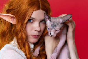 portrait of cosplay elf young redhead woman with sphynx kitten on red background